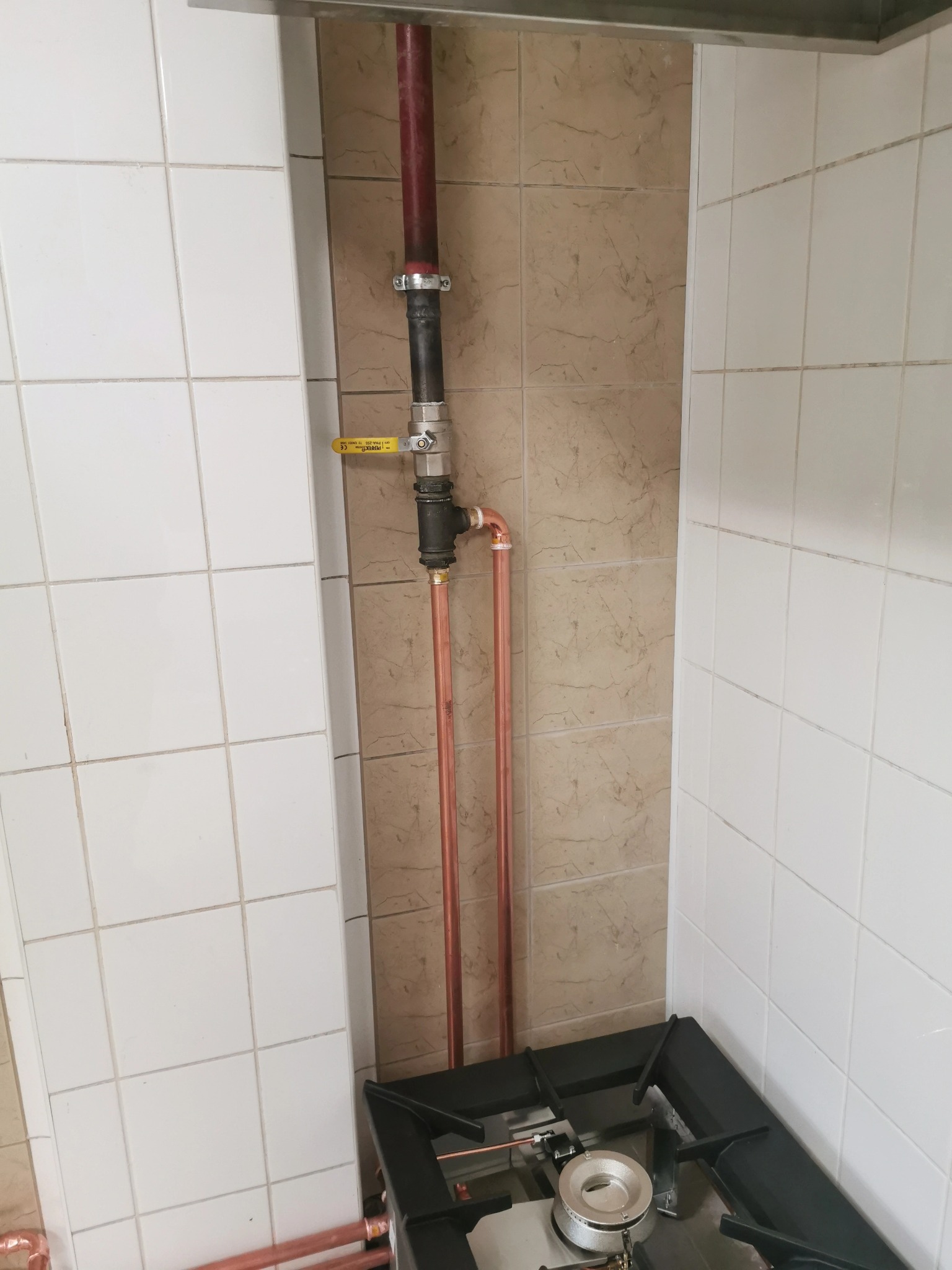 Nowe miedziane rury gazowe z zaworem odcinającym, połączone z istniejącą instalacją w kuchni z płytkami w dwóch odcieniach beżu, tuż obok kuchenki gazowej.