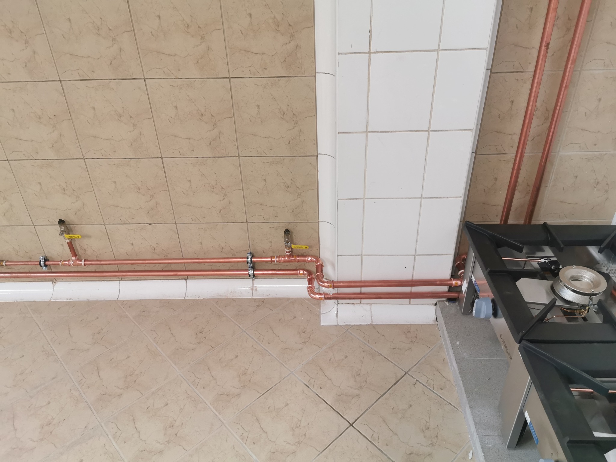 Miedziane rury gazowe z zaworami, zamontowane pod sufitem w kuchni z płytkami, obok kuchenki gazowej, widok z góry.