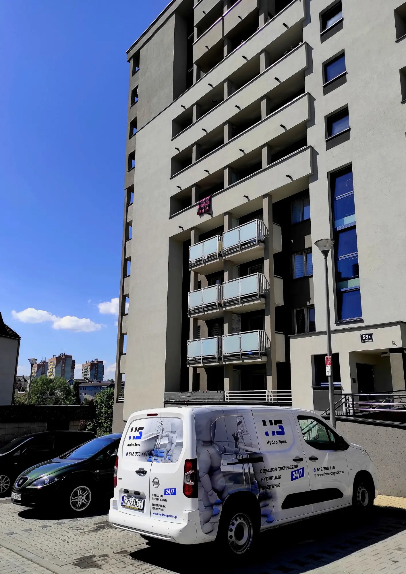 Samochód serwisowy z logo firmy Hydro Spec zaparkowany przed nowoczesnym budynkiem mieszkalnym z balkonami.