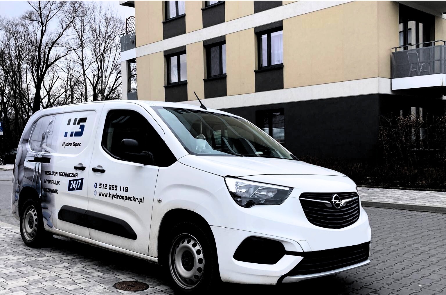 Biały samochód dostawczy firmy Hydro Spec z numerem telefonu i adresem strony internetowej na tle budynku mieszkalnego.