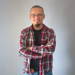 Uśmiechnięty mężczyzna w okularach, koszuli w kratę i jeansach, z miarką budowlaną przy pasku, stojący na tle szarej ściany.