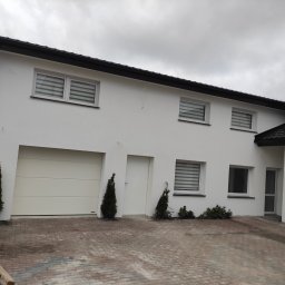 Piotr Stetsiuk - Jasny dom jednorodzinny w Gdańsku z białą elewacją i grafitowymi akcentami na dachu i oknach, widok z zewnątrz na świeżo położoną kostkę brukową. Na pierwszym planie paleta z materiałami budowlanymi.