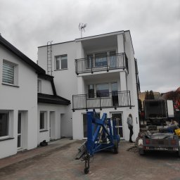 Piotr Stetsiuk - Dwupiętrowy budynek mieszkalny z białymi ścianami i czarnymi balkonami, drabina na elewacji, niebieski podnośnik koszowy i przyczepa na brukowanym podwórku, pochmurne niebo.