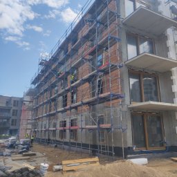 Piotr Stetsiuk - Budynek wielorodzinny w trakcie prac elewacyjnych, widoczne rusztowania, cegły, częściowo otynkowana ściana, balkony, materiały budowlane na ziemi i dwóch pracowników w odblaskowych kamizelkach.