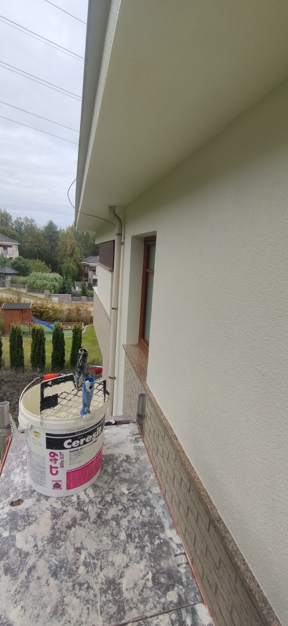 Pomalowana na biało elewacja domu jednorodzinnego, widoczny fragment okna z brązową ramą, wiadro z farbą marki Ceresit CT 49 i narzędzia malarskie na rusztowaniu, w tle zieleń ogrodu i inne budynki.