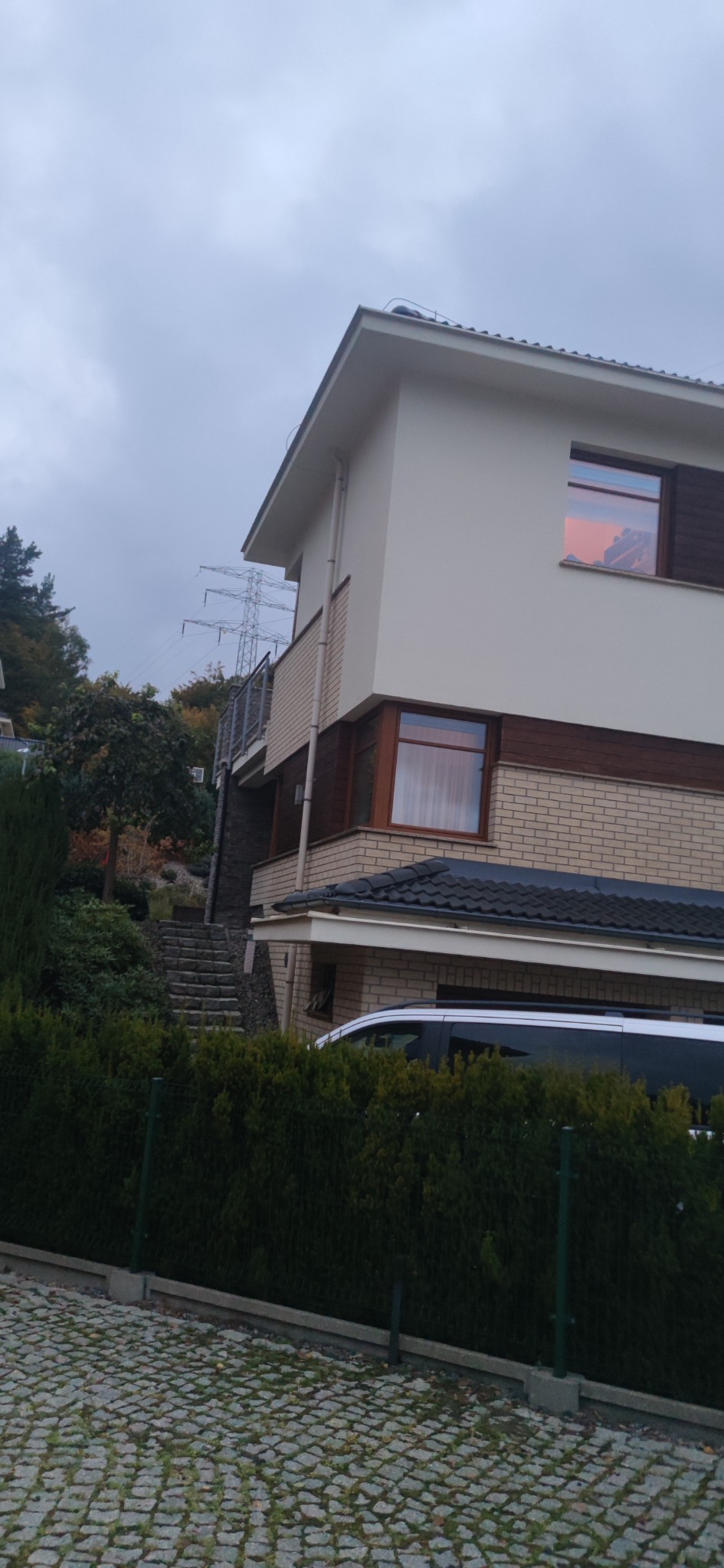 Dwukondygnacyjny dom z jasną elewacją, ceglanym wykończeniem i drewnianymi oknami, widziany z poziomu ulicy w pochmurny dzień.