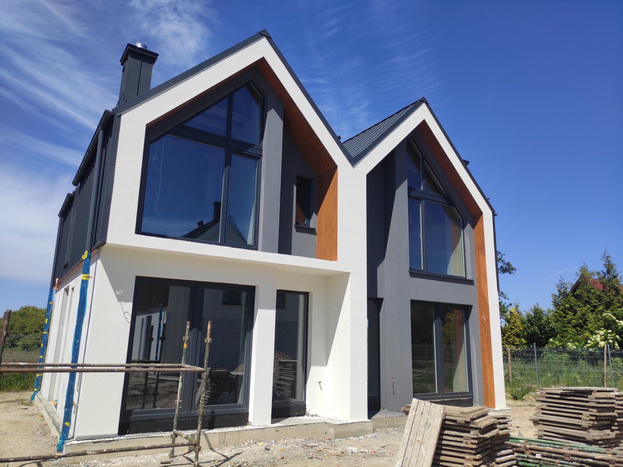 Nowoczesny dom jednorodzinny z dwuspadowym dachem, pomalowany na szaro i biało, z dużymi oknami, widoczne rusztowania i materiały budowlane sugerujące prace wykończeniowe elewacji.