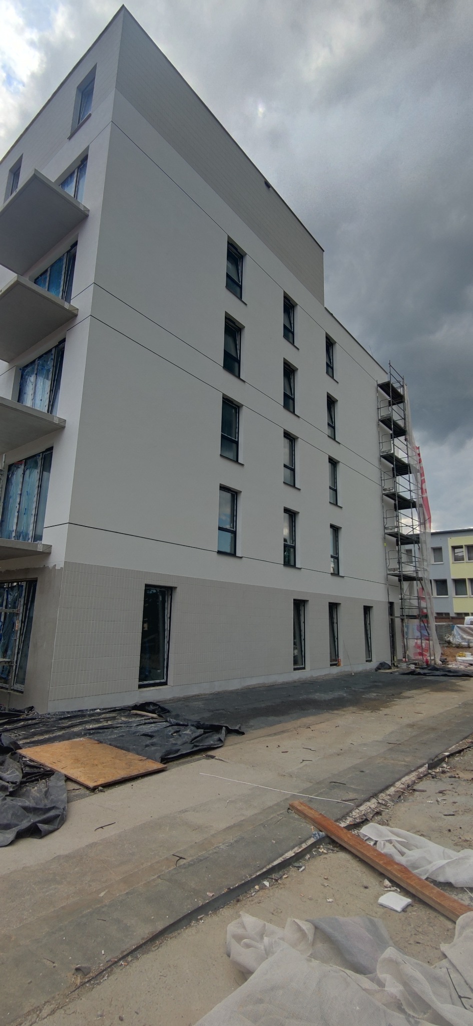 Wielopiętrowy budynek z częściowo wykończoną elewacją w odcieniach bieli i szarości, widoczne rusztowania i zabezpieczenia okien folią, teren budowy w nieładzie.