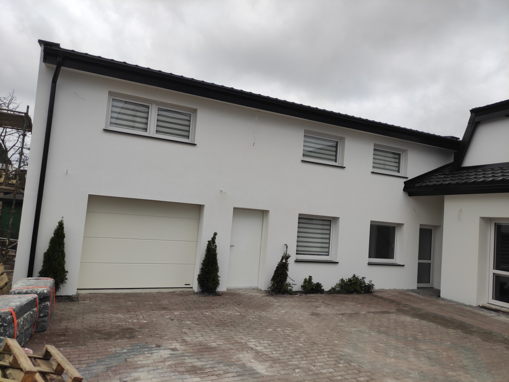 Jasny dom jednorodzinny w Gdańsku z białą elewacją i grafitowymi akcentami na dachu i oknach, widok z zewnątrz na świeżo położoną kostkę brukową. Na pierwszym planie paleta z materiałami budowlanymi.