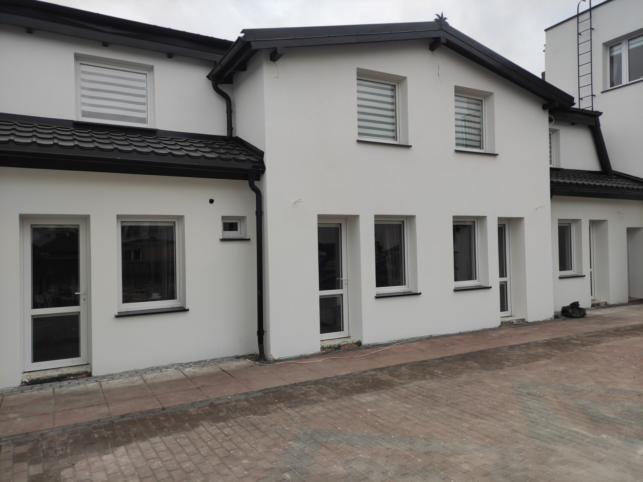 Świeżo otynkowana i pomalowana na biało elewacja dwukondygnacyjnego domu z czarnym dachem i rynnami, widok z poziomu kostki brukowej.