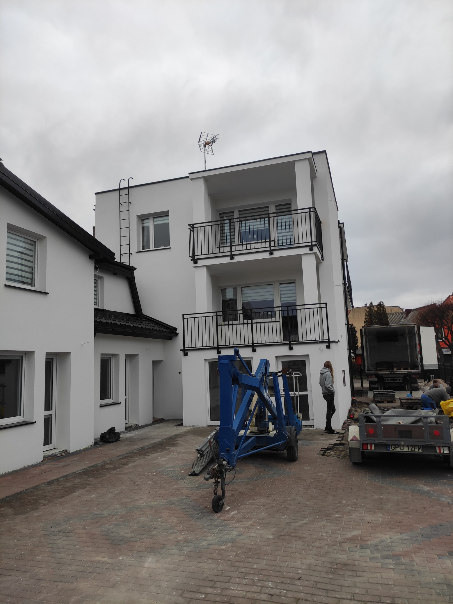 Dwupiętrowy budynek mieszkalny z białymi ścianami i czarnymi balkonami, drabina na elewacji, niebieski podnośnik koszowy i przyczepa na brukowanym podwórku, pochmurne niebo.