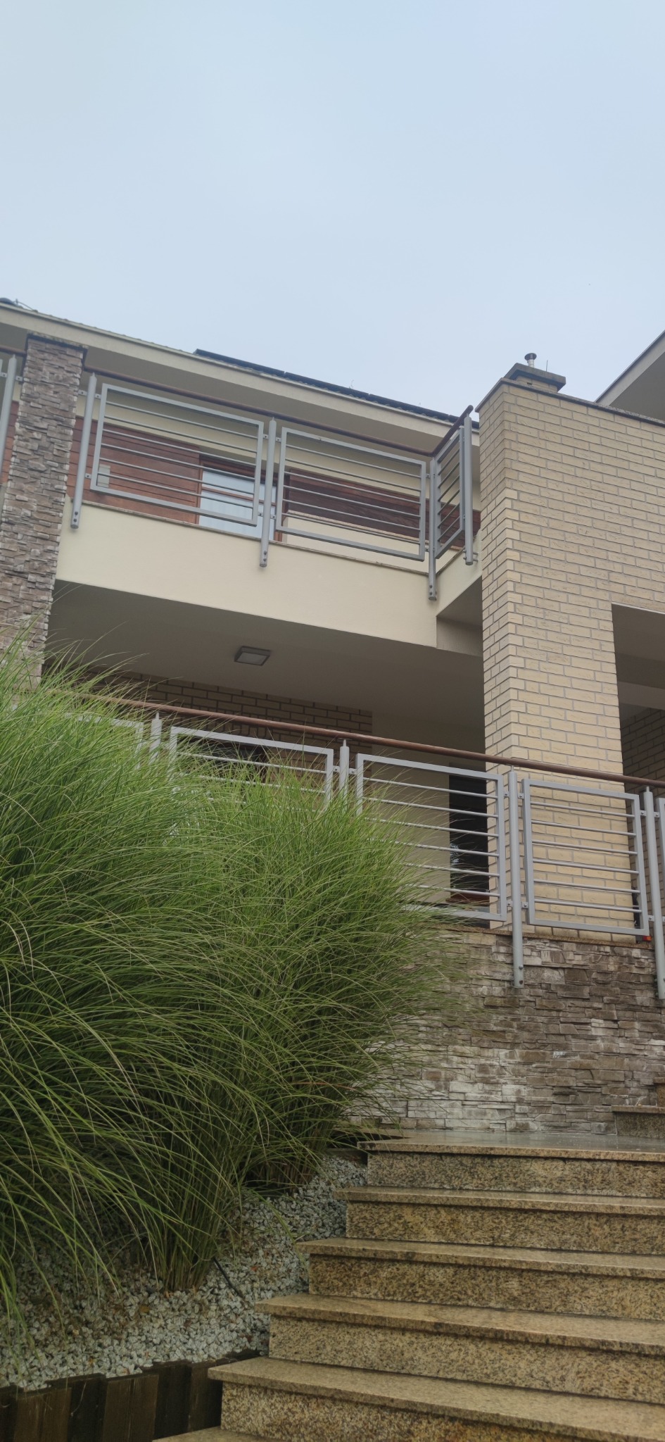 Dwupiętrowy dom z elementami elewacji z cegły i kamienia, widok z dołu na balkon z metalową balustradą i schody wejściowe z piaskowca, przysłonięte ozdobną trawą.