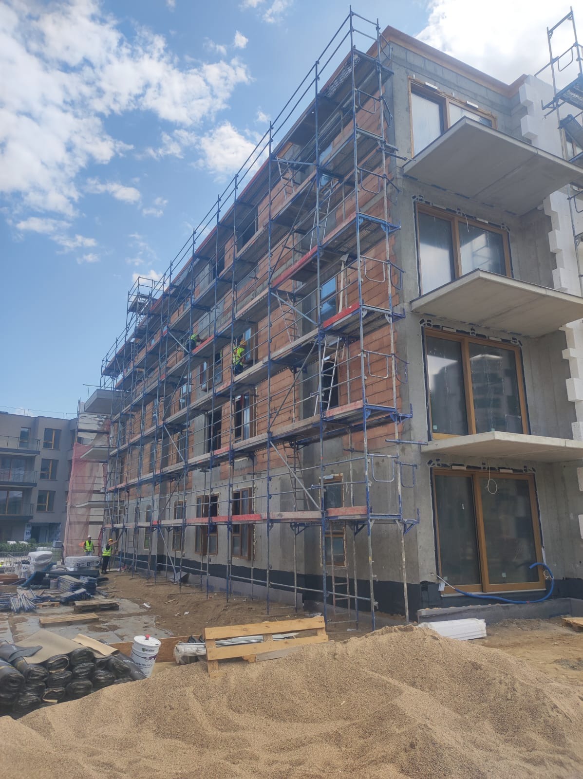 Budynek wielorodzinny w trakcie prac elewacyjnych, widoczne rusztowania, cegły, częściowo otynkowana ściana, balkony, materiały budowlane na ziemi i dwóch pracowników w odblaskowych kamizelkach.
