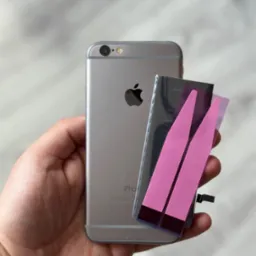 Wymiana baterii w telefonie iPhone w Rybniku: dłoń trzyma srebrny telefon iPhone 6s obok nowej baterii z różowymi paskami zabezpieczającymi klej.