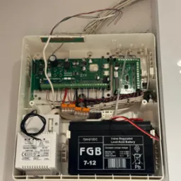 Otwarta skrzynka centrali alarmowej z widoczną płytą główną, transformatorem, akumulatorem i licznymi przewodami. Pomarańczowe złączki Wago na przewodach. Widoczne oznaczenia na akumulatorze...