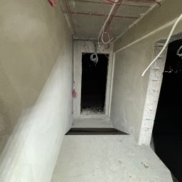 LANRUNNER Mateusz Sałek - Surowy stan wnętrza w trakcie remontu, widoczne kable elektryczne wystające z sufitu i ścian, betonowe ściany w trakcie tynkowania, ciemne wejście do kolejnego pomieszczenia.
