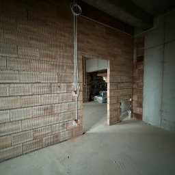 LANRUNNER Mateusz Sałek - Surowy stan wnętrza w Tarnobrzegu: widoczne przewody elektryczne na ceglanej ścianie, surowy betonowy strop i podłoga, w głębi pomieszczenia worki z materiałami budowlanymi.