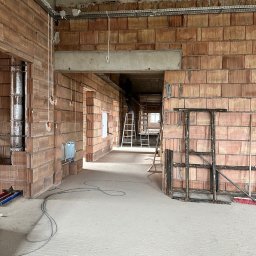 LANRUNNER Mateusz Sałek - Wnętrze budynku w trakcie budowy, ściany z czerwonej cegły, widoczne instalacje elektryczne w korytach, przewody, drabiny w głębi, surowy betonowy strop.