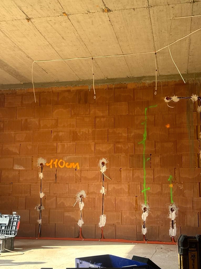 Surowa ściana z cegły z widocznymi przewodami elektrycznymi w trakcie montażu, zaznaczone pionowe linie laserowe i wysokość 110cm, sufit betonowy, narzędzia w tle.