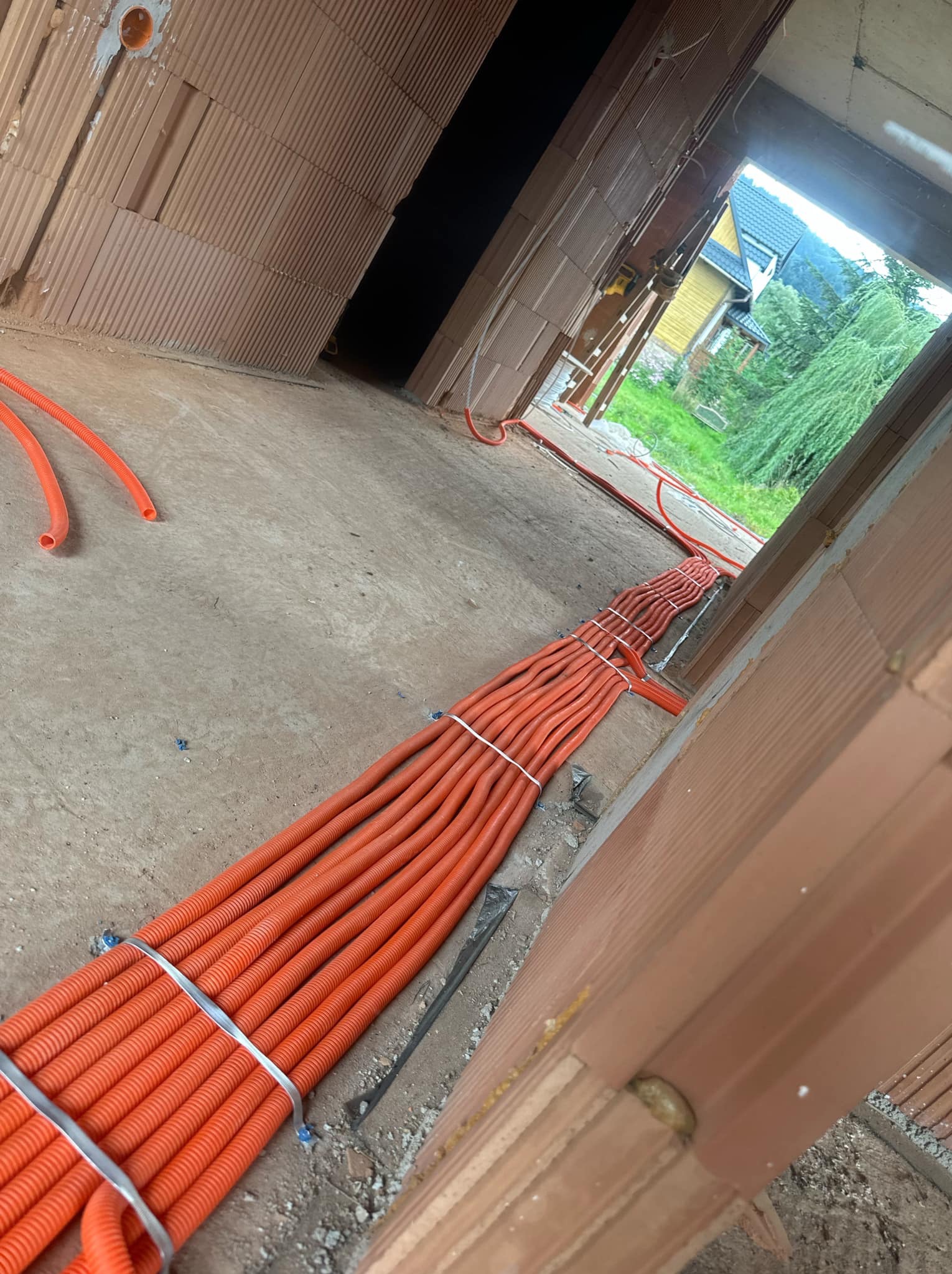 Pomarańczowe rury instalacyjne ułożone równolegle na podłodze w surowym stanie budynku, widoczne ściany z cegieł i otwór drzwiowy z widokiem na zieleń.