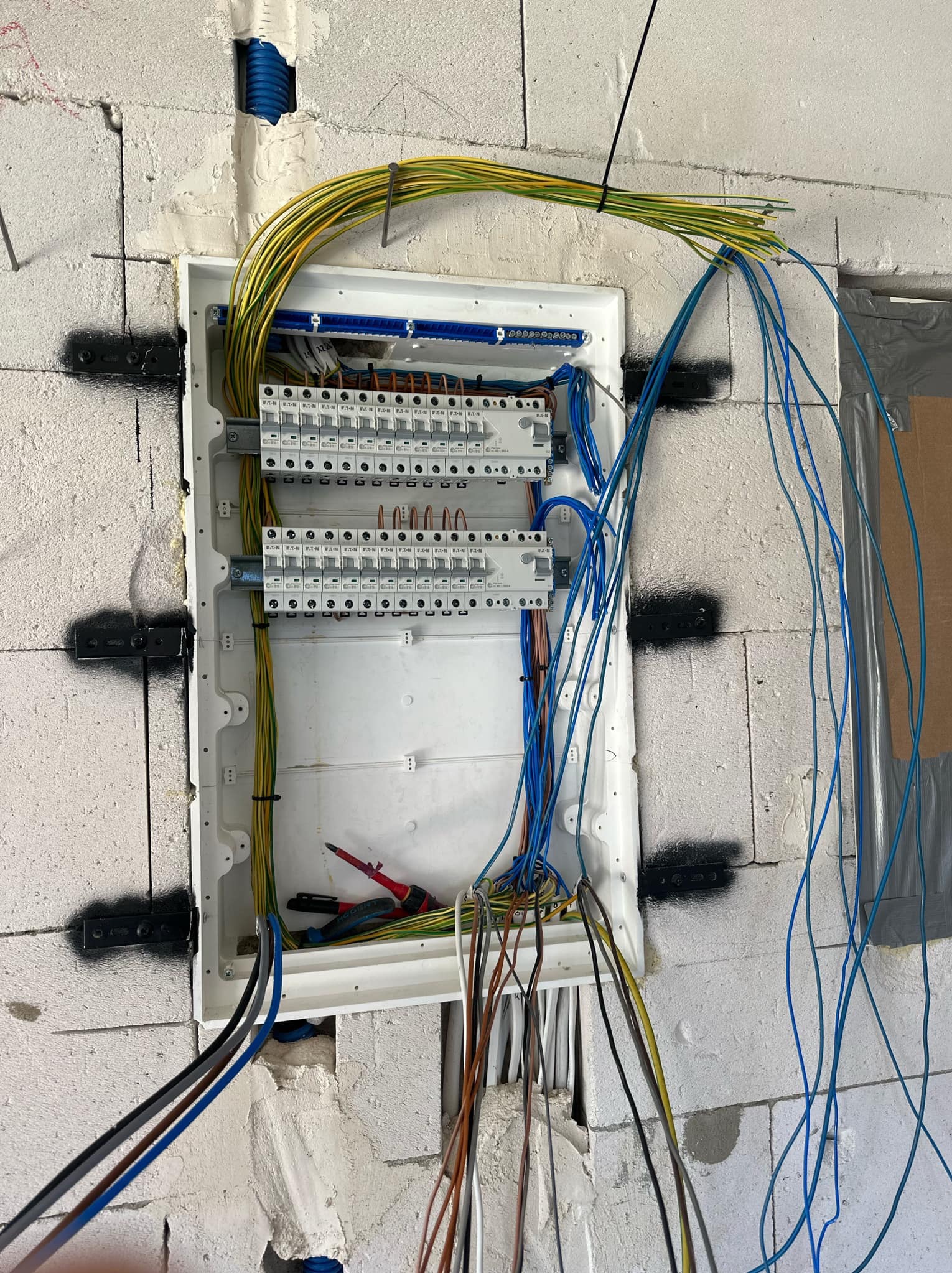 Otwarta skrzynka rozdzielcza z podłączonymi przewodami w surowym wnętrzu, widoczne narzędzia wewnątrz i fragment ściany z betonu komórkowego.
