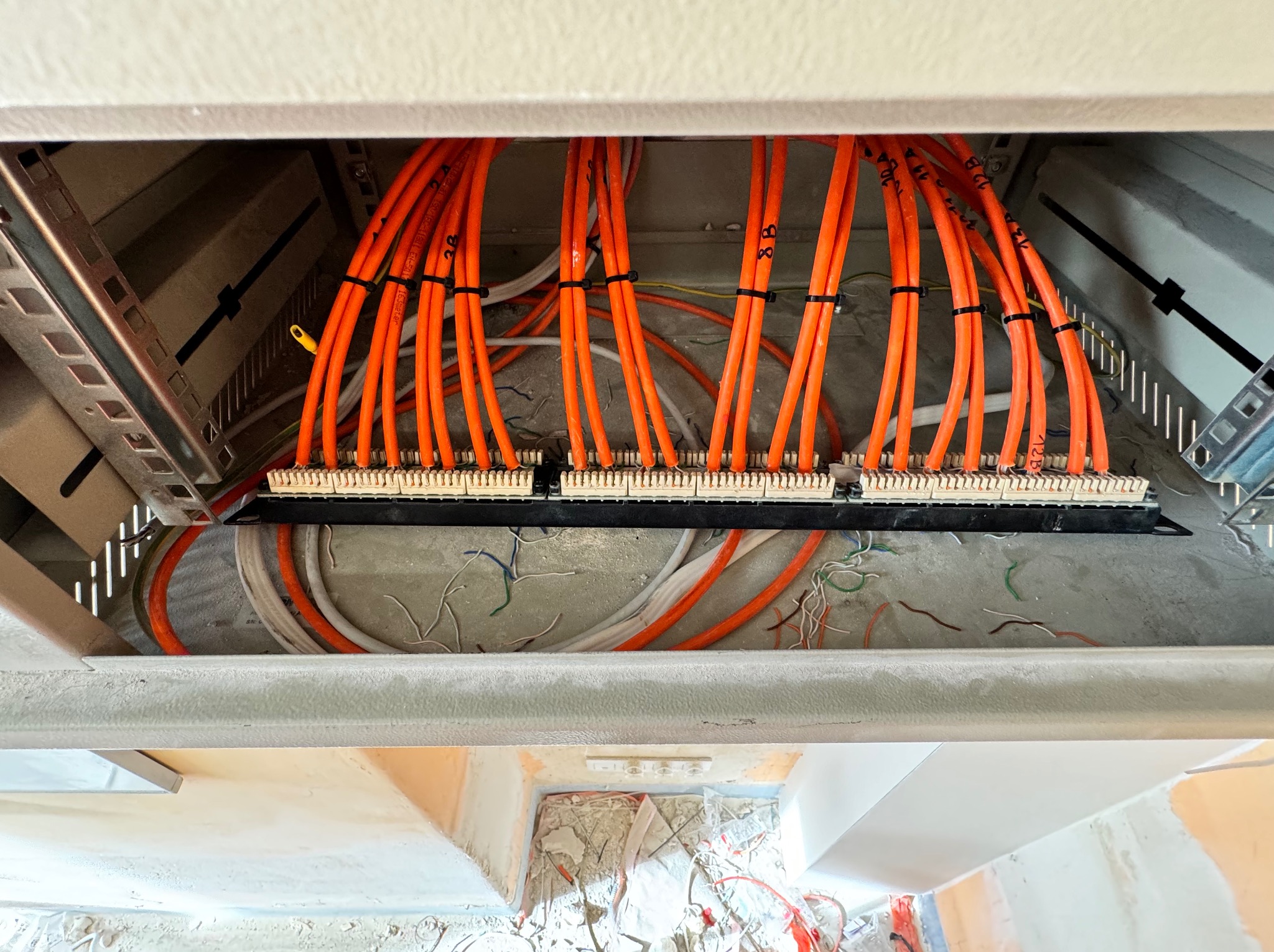 Pomarańczowe kable sieciowe uporządkowane w szafie rack, oznaczone czarnymi numerami, widoczne pozostałości po pracach budowlanych i inne kable w tle.