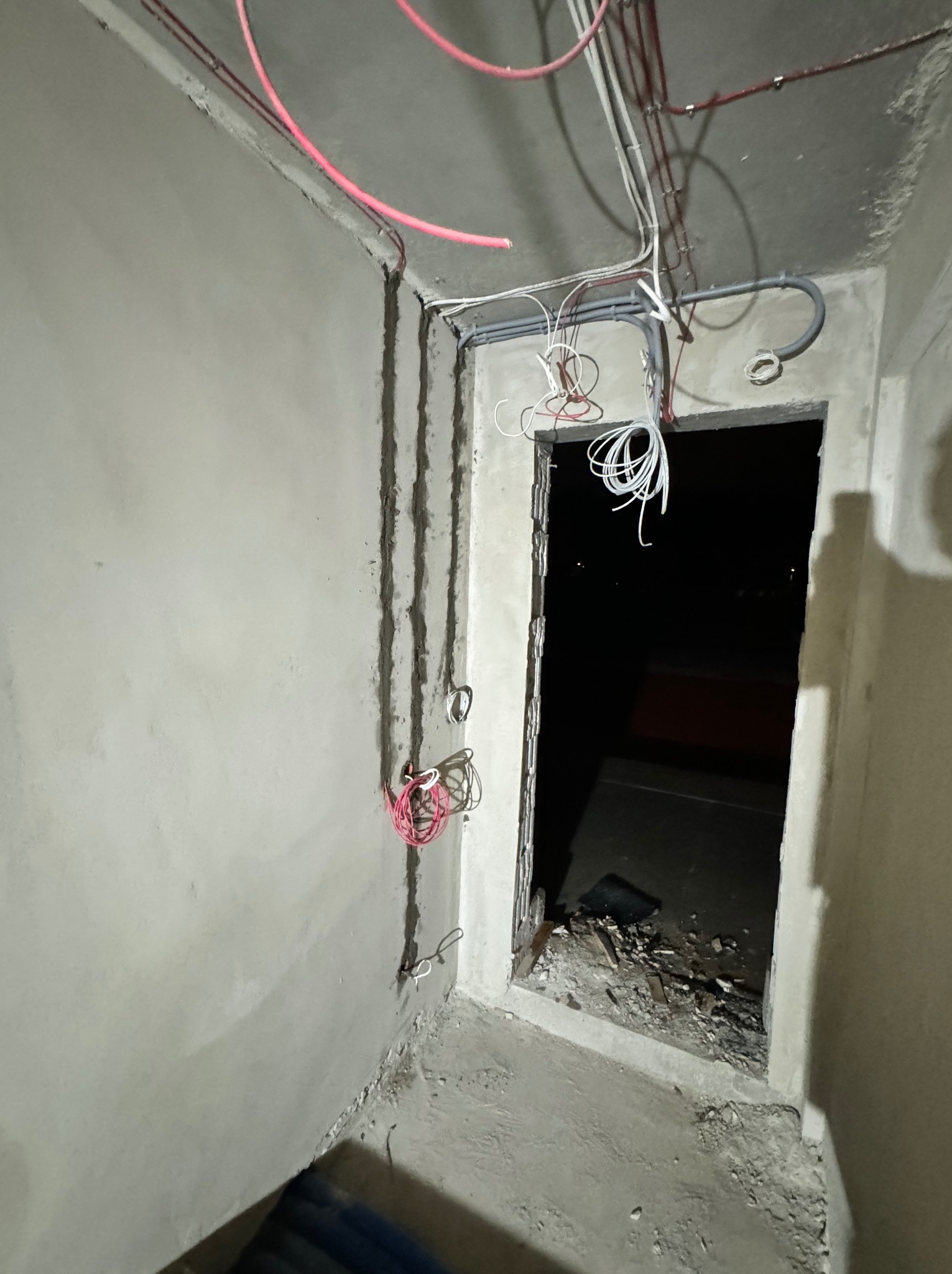 Surowy stan wnętrza w trakcie prac: widoczne kable elektryczne prowadzone po suficie i ścianach, bruzdy w ścianach, nieotynkowany otwór drzwiowy z wystającymi przewodami.