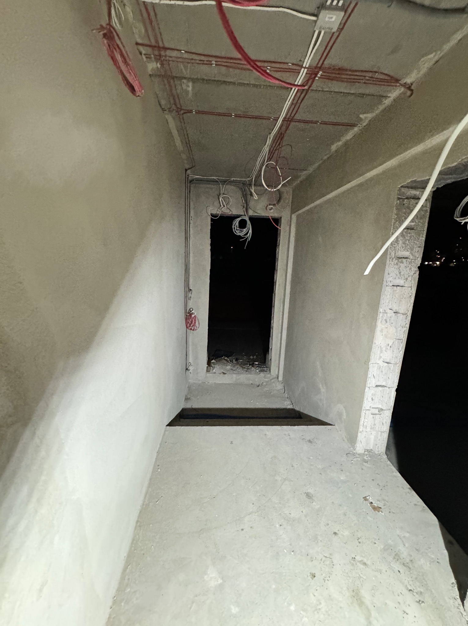 Surowy stan wnętrza w trakcie remontu, widoczne kable elektryczne wystające z sufitu i ścian, betonowe ściany w trakcie tynkowania, ciemne wejście do kolejnego pomieszczenia.