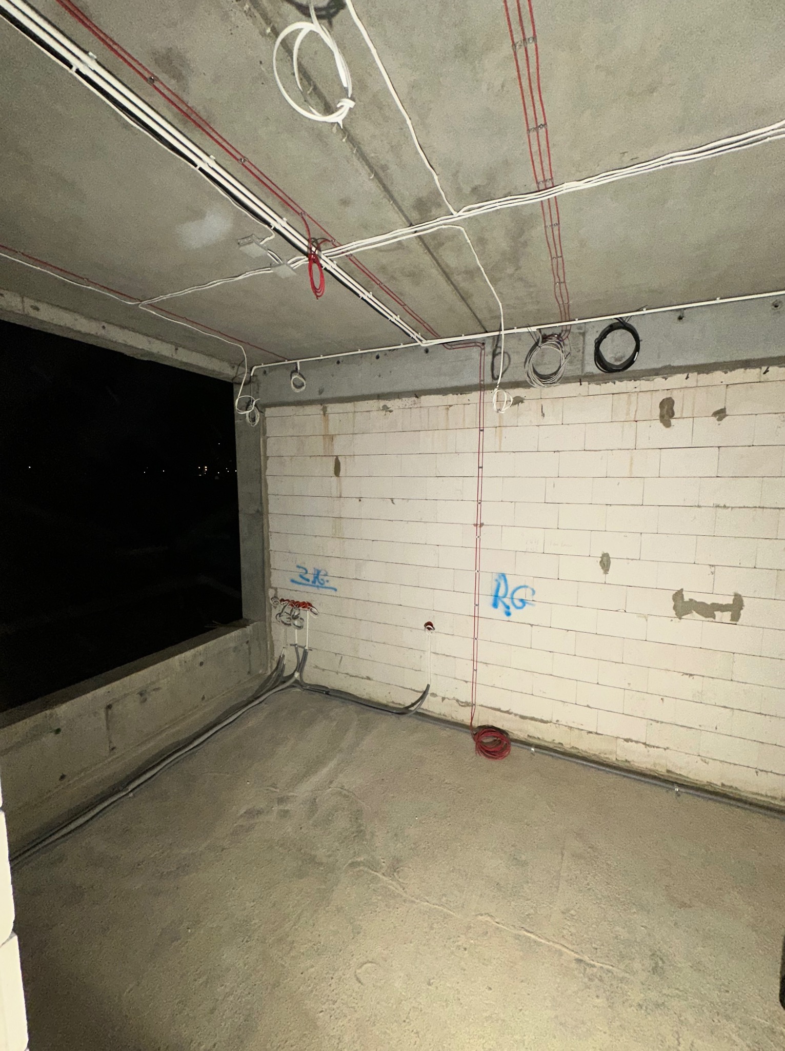Surowy stan wnętrza w trakcie prac elektrycznych: widoczne kable poprowadzone po suficie i ścianach, ściany z białego bloczka z surowym tynkiem, betonowa podłoga, okno z widokiem na noc.