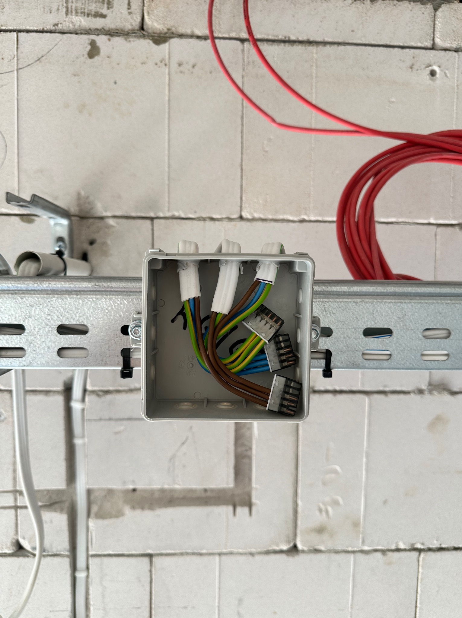 Szara puszka instalacyjna zamontowana na metalowej szynie w surowym wnętrzu z bloczków, widoczne podłączone przewody w kolorach: brązowym, niebieskim, zielono-żółtym oraz czerwony kabel zwinięty...