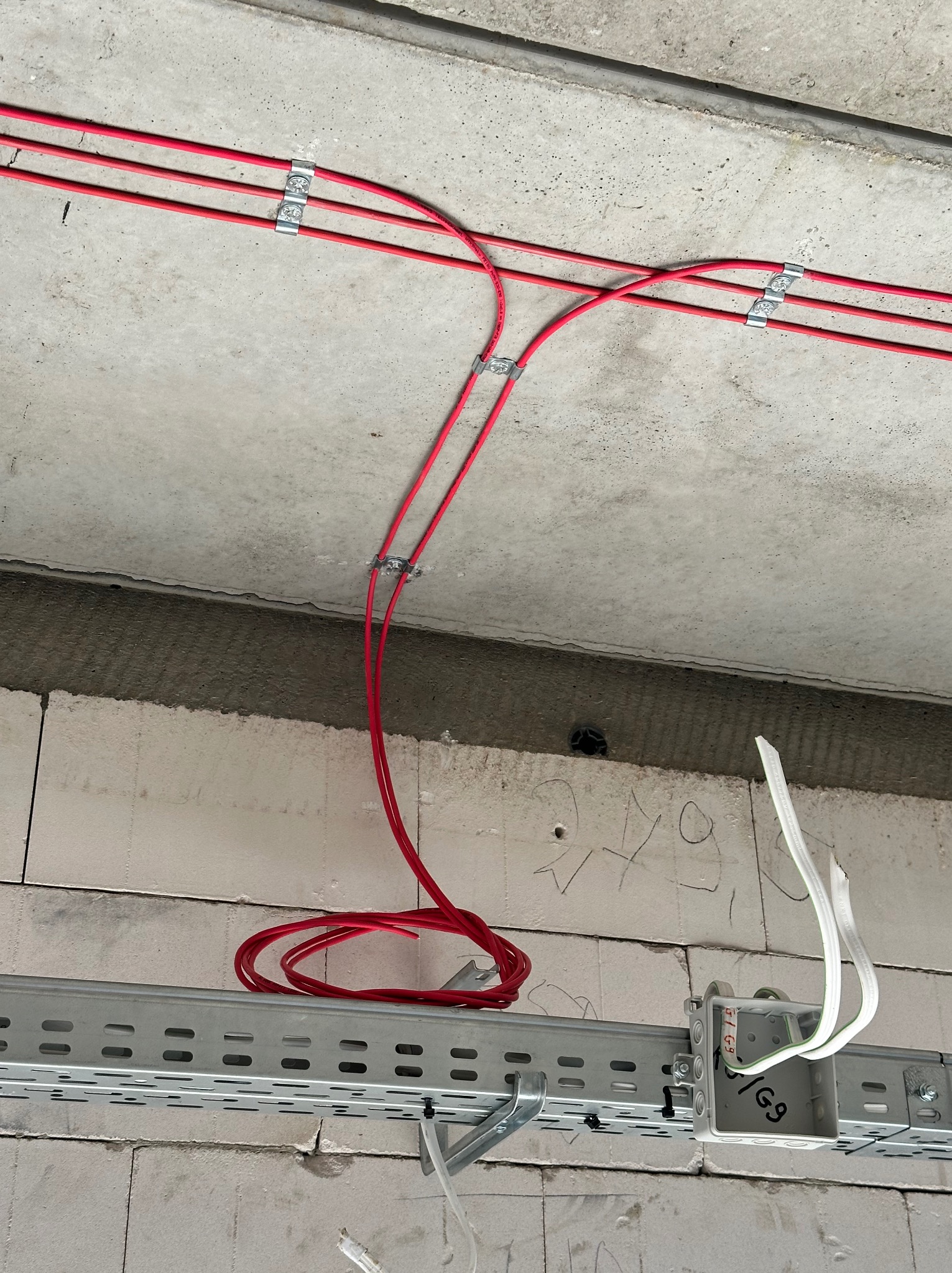 Czerwone kable elektryczne zamocowane do sufitu za pomocą metalowych uchwytów, z widoczną puszką instalacyjną i szyną montażową na ścianie z białych bloczków.