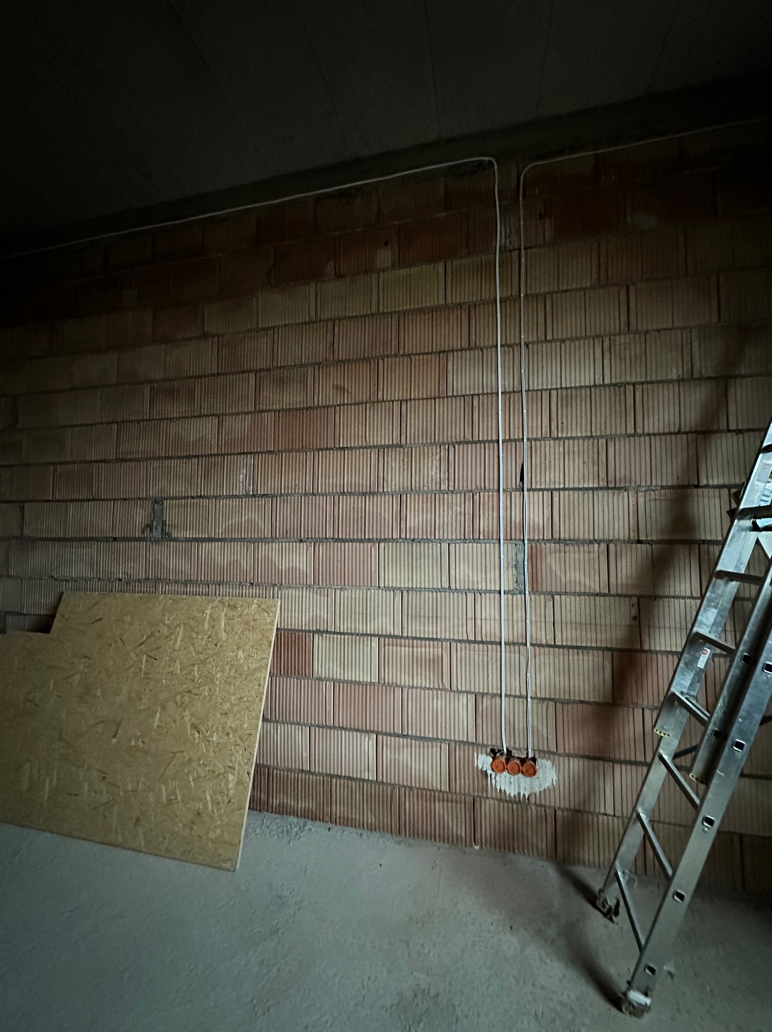 Surowa ściana z cegły dziurawki z poprowadzonymi przewodami elektrycznymi i puszkami pod gniazdka, obok drabina aluminiowa, na podłodze płyta OSB.