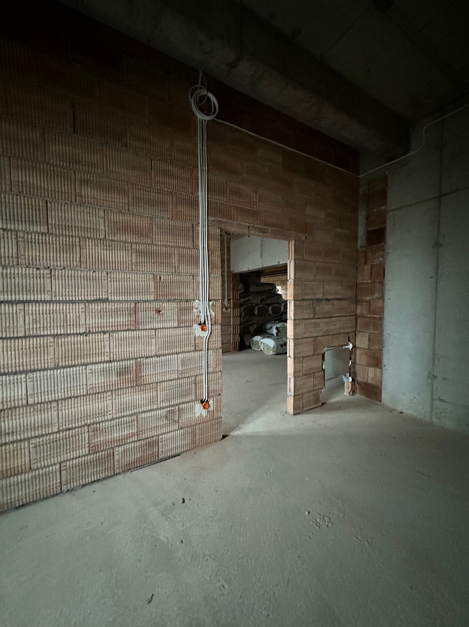 Surowy stan wnętrza w Tarnobrzegu: widoczne przewody elektryczne na ceglanej ścianie, surowy betonowy strop i podłoga, w głębi pomieszczenia worki z materiałami budowlanymi.