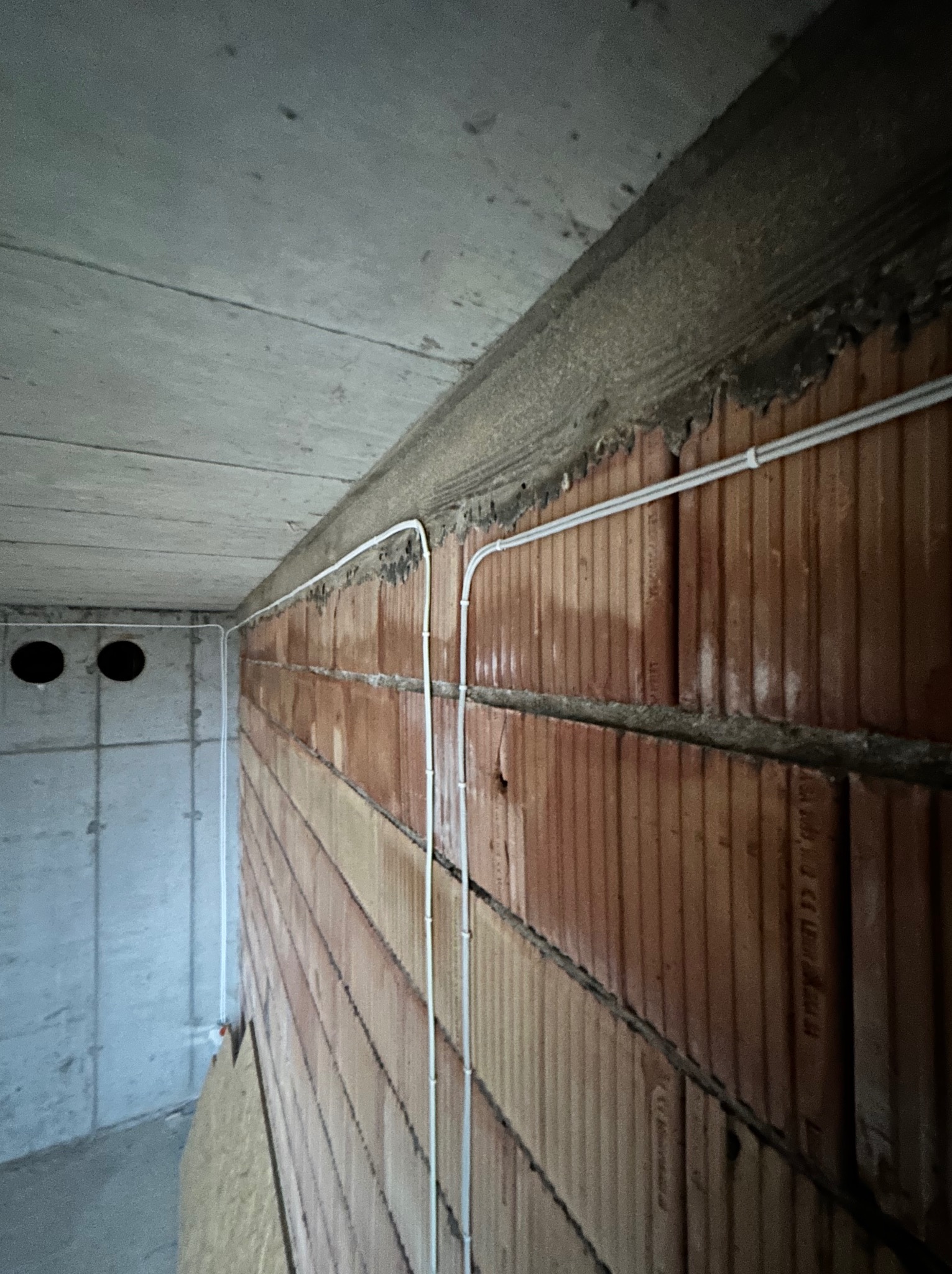 Instalacja elektryczna: białe przewody ułożone wzdłuż ceglanej ściany pod betonowym sufitem w surowym stanie.