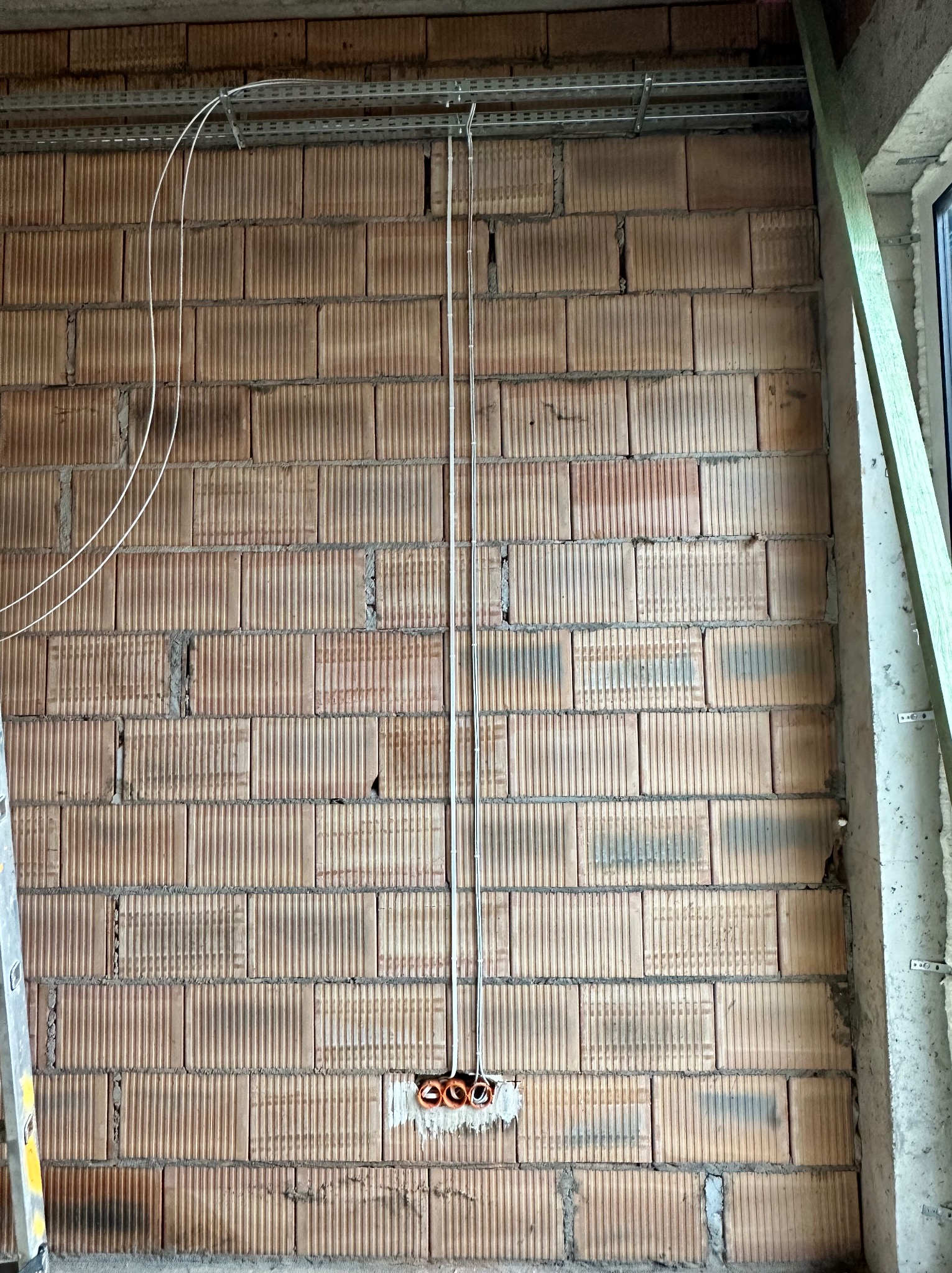 Ściana z pustaków z instalacją elektryczną w trakcie montażu: kable prowadzone w korytkach i rurkach, puszka elektryczna z pomarańczowymi peszlami w ścianie.