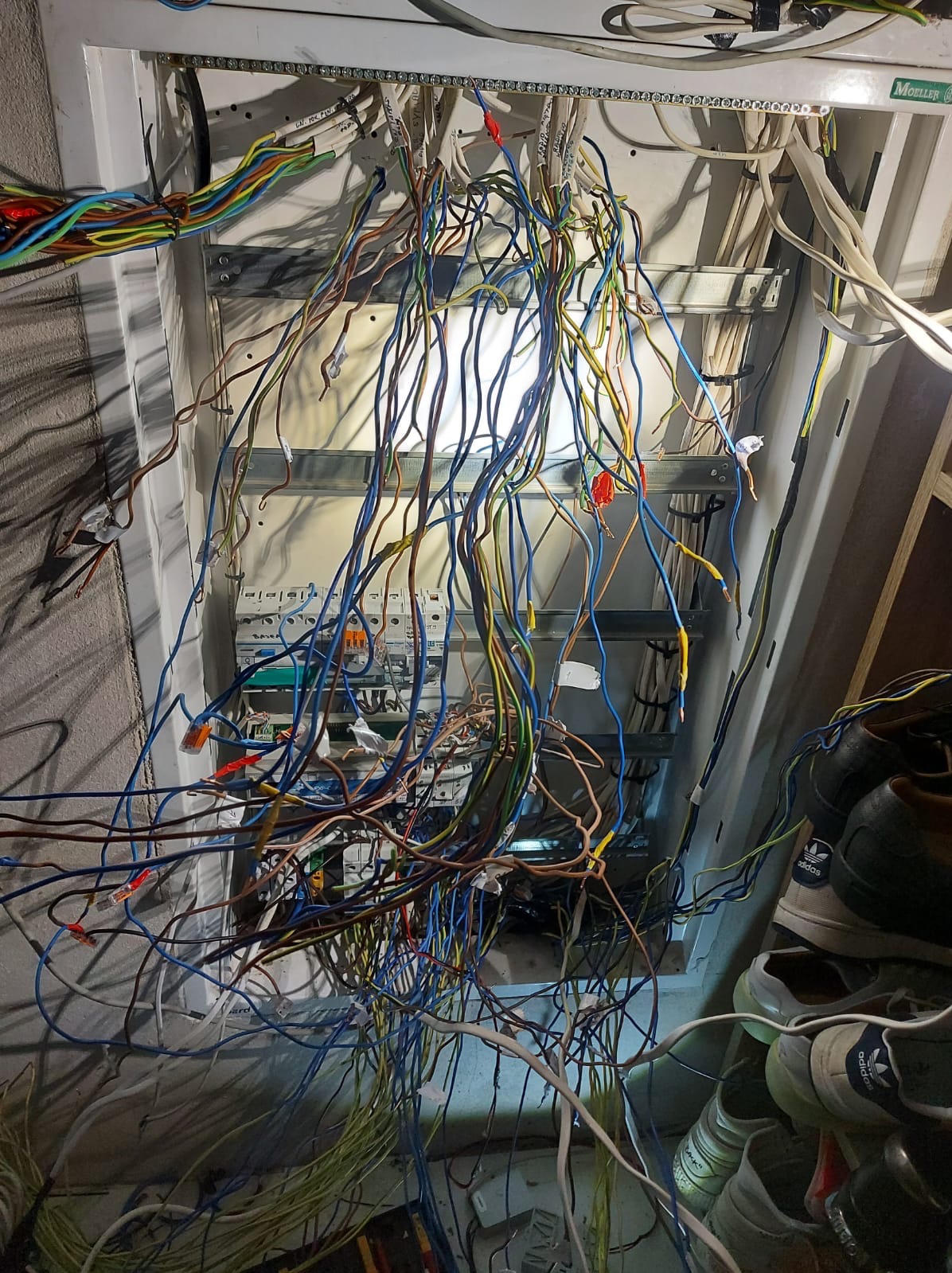 Otwarta skrzynka elektryczna z licznymi kablami w różnych kolorach, widoczne elementy instalacji i złącza, chaotyczny układ przewodów, oświetlenie robocze wewnątrz skrzynki, buty w pobliżu.