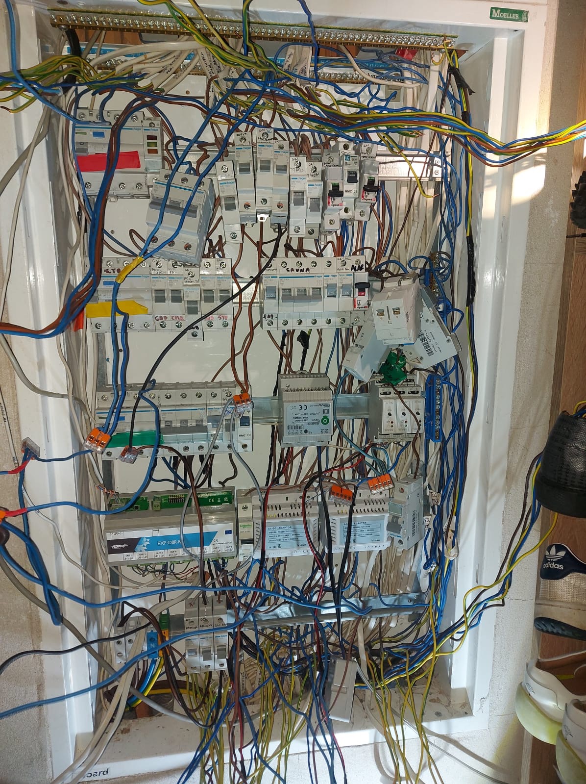 Otwarta skrzynka elektryczna z licznymi przewodami w kolorach niebieskim, żółtym i brązowym, widoczne zabezpieczenia i moduły, częściowo zasłonięta butami w prawym dolnym rogu, etykieta 'Poprawka'.