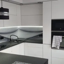 Nowoczesna kuchnia z białymi szafkami, czarnym blatem i czarno-srebrnymi lampami wiszącymi, z abstrakcyjnym wzorem na ścianie nad blatem.