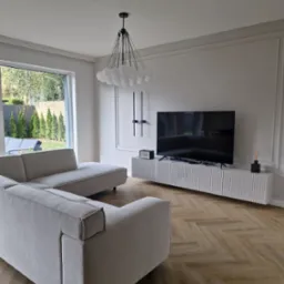 Jasny salon z narożną sofą, telewizorem na szafce RTV z lamelami, parkietem w jodełkę i lampą w kształcie białych kul.