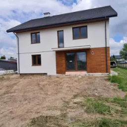 Dwukondygnacyjny dom z jasną elewacją i drewnianym wykończeniem dolnej części, widoczny fragment dachu z czarną dachówką, teren wokół domu częściowo zagospodarowany, błękitne niebo z obłokami w tle.