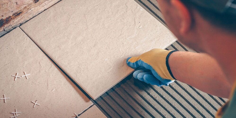 Beżowa płytka ceramiczna układana na podłodze, widoczne krzyżyki dystansowe i warstwa kleju z rowkami, ręka w roboczej rękawicy dociska krawędź.