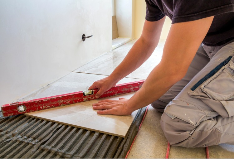 Pracownik w spodniach roboczych, używając poziomicy, układa jasną, marmurkową płytkę na warstwie kleju na podłodze w pomieszczeniu.