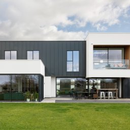 SQ LINE SPÓŁKA Z OGRANICZONĄ ODPOWIEDZIALNOŚCIĄ - Nowoczesny dom z dużymi przesuwnymi oknami i szarym metalowym sidingiem, widok na ogród z meblami ogrodowymi i grillem.