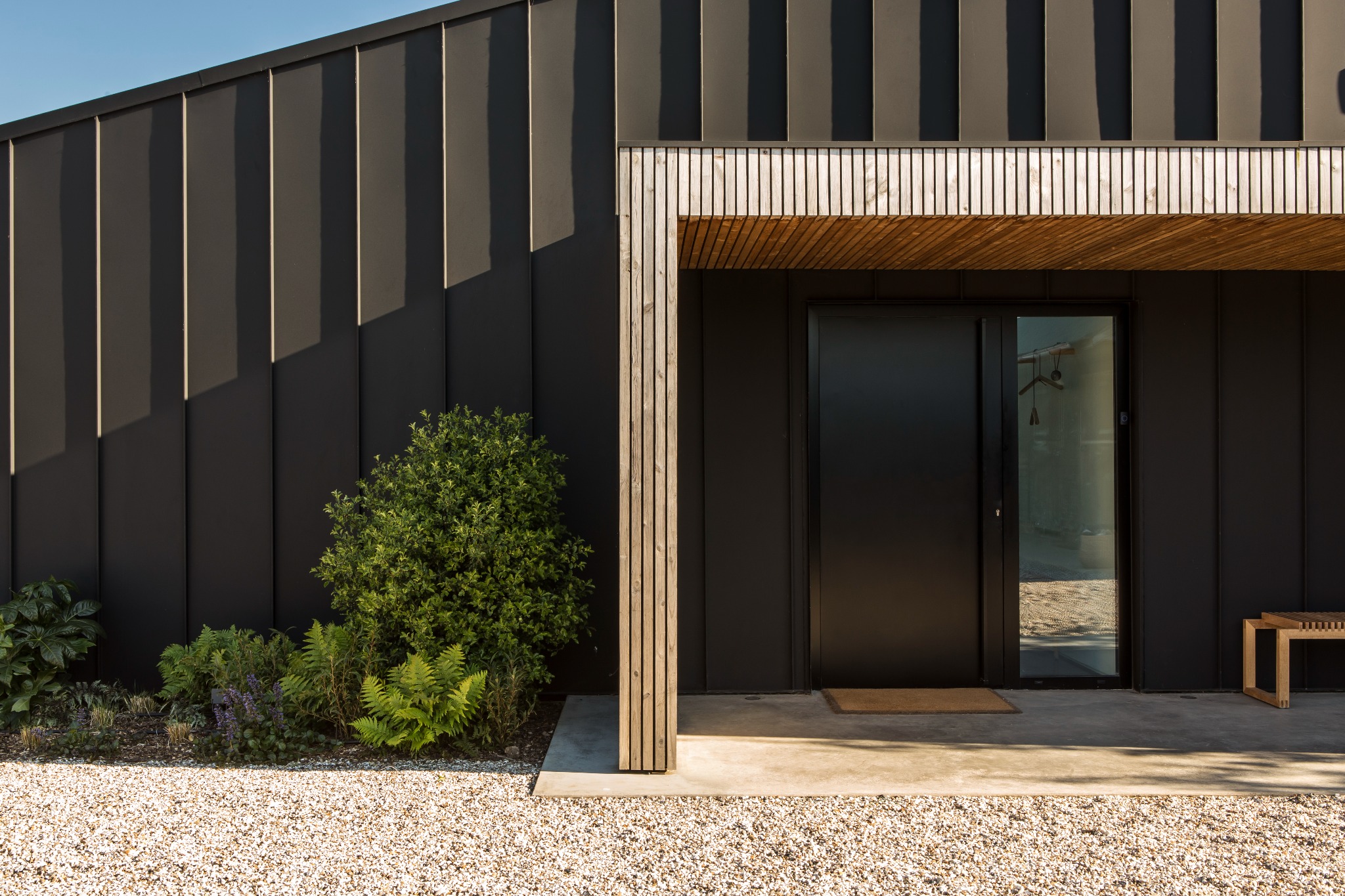 Nowoczesny dom z czarną elewacją z blachy, drewnianym zadaszeniem nad wejściem z przesuwnymi drzwiami i minimalistycznym ogrodem z krzewami i żwirem.