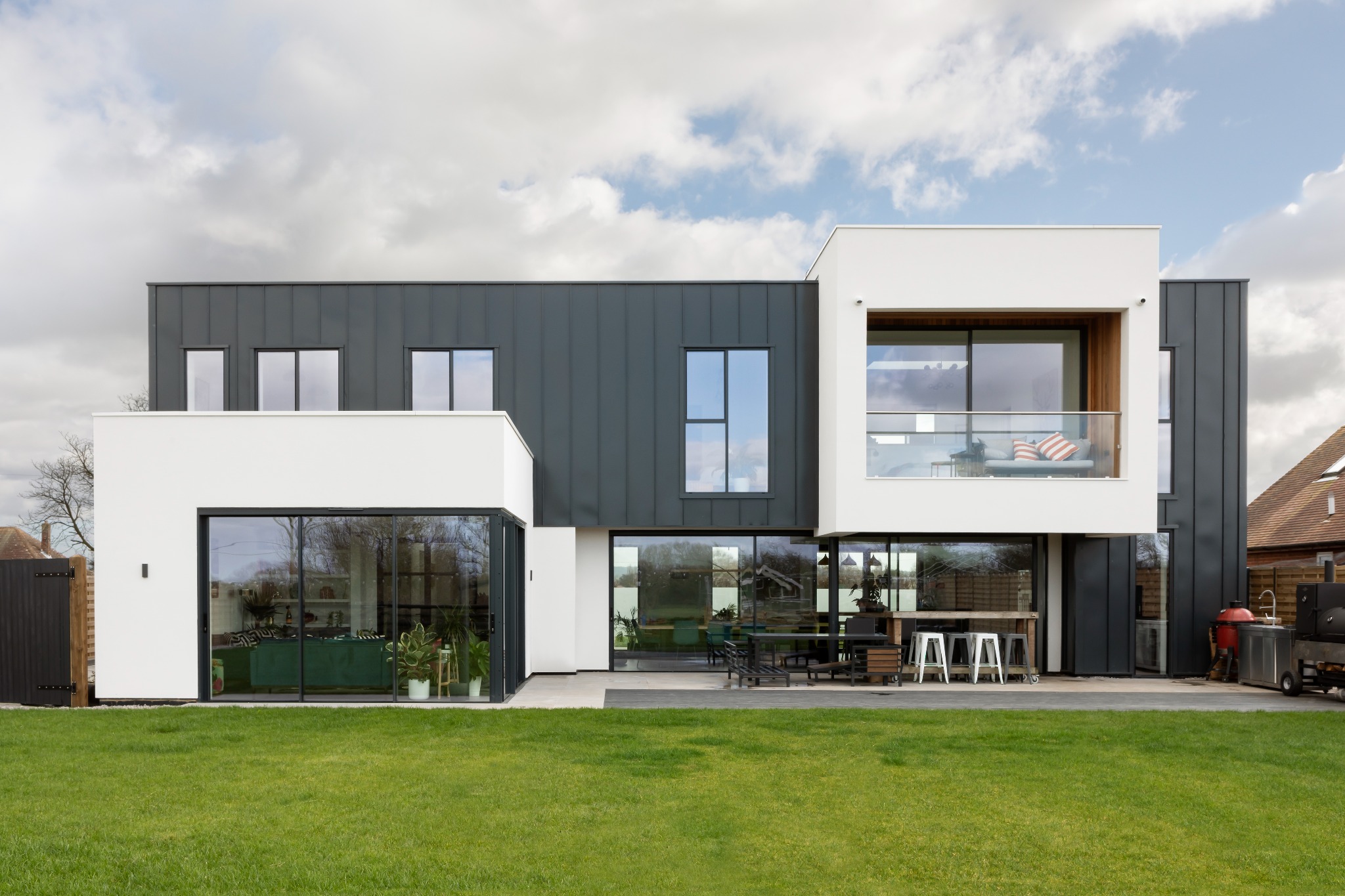 Nowoczesny dom z dużymi przesuwnymi oknami i szarym metalowym sidingiem, widok na ogród z meblami ogrodowymi i grillem.