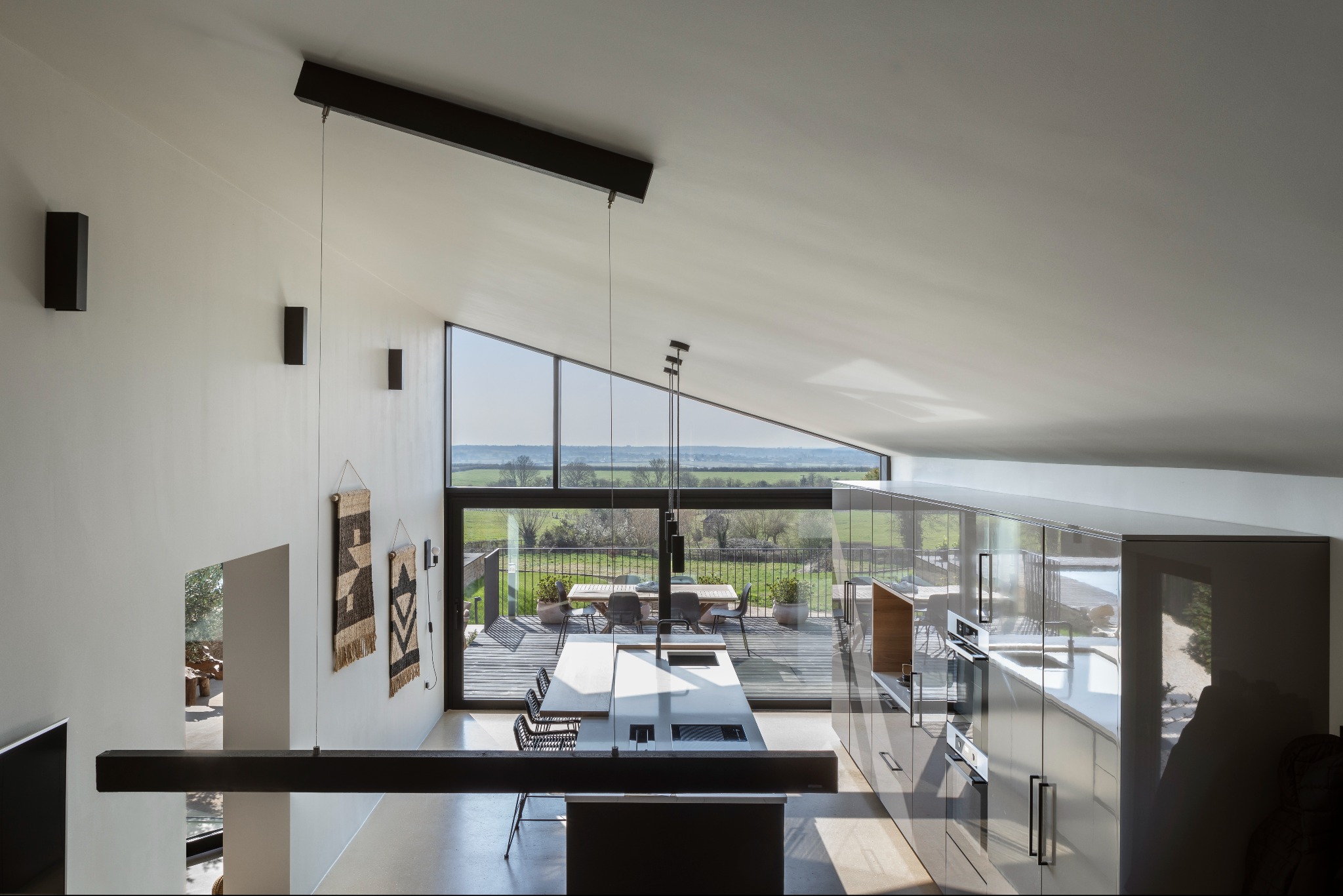 Wnętrze nowoczesnego domu z dużymi oknami wychodzącymi na taras i zielony krajobraz, widok z góry na minimalistyczną kuchnię z błyszczącymi szafkami i wyspą, dekoracyjne tkaniny na ścianie.