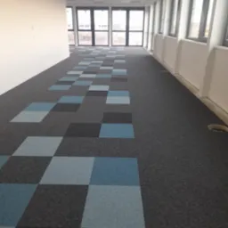 Wykładzina dywanowa w biurze z wzorem szachownicy z szarych i niebieskich kwadratów, widok z perspektywy podłogi w kierunku przeszklonych drzwi wejściowych.