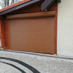 Zamknięta brązowa roleta garażowa w drewnianej obudowie, widoczny fragment elewacji budynku i wzorzysty podjazd z kostki brukowej.