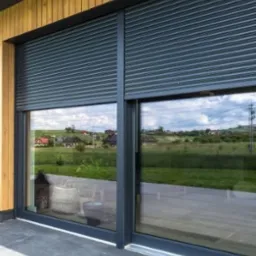 Zewnętrzna, szara roleta częściowo opuszczona na oknie tarasowym z widokiem na zielony krajobraz i zabudowania w tle.