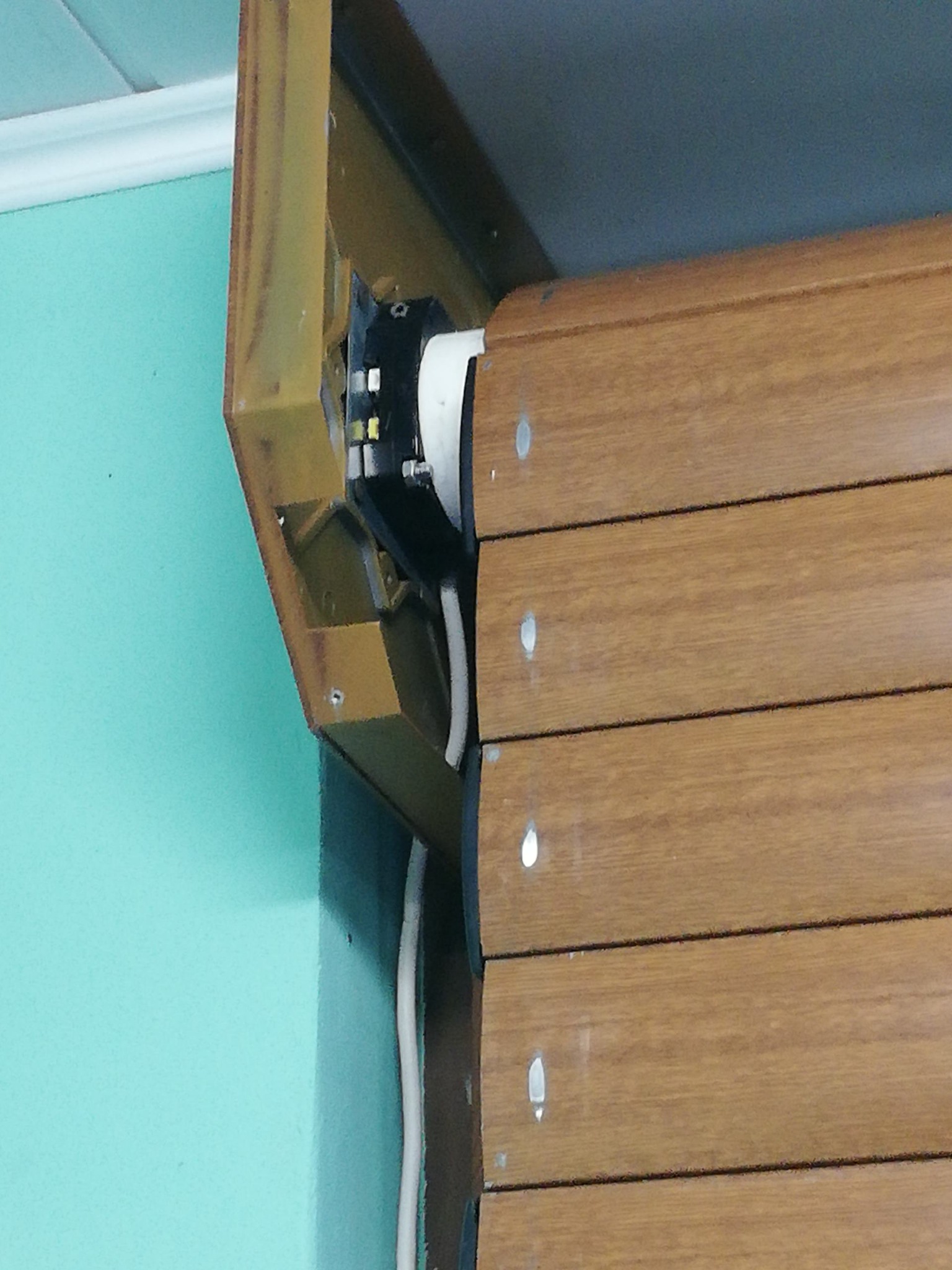 Fragment montażu rolety zewnętrznej z widoczną mechaniką i okablowaniem, imitacja drewna, tło w odcieniach turkusu i szarości.